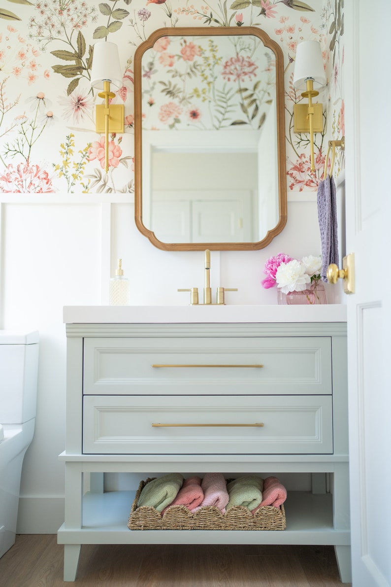 May include: A light green bathroom vanity with two drawers and a lower shelf. The vanity has gold hardware and a white countertop. A mirror with a wood frame hangs above the vanity. The wall behind the vanity is covered in floral wallpaper.