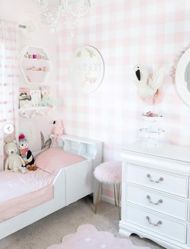 A cute and cozy girl's bedroom with pink gingham peel-and-stick wallpaper, creating a soft, playful backdrop. The room features a white toddler bed, plush toys, a white dresser, decorative shelves, and a swan wall mount