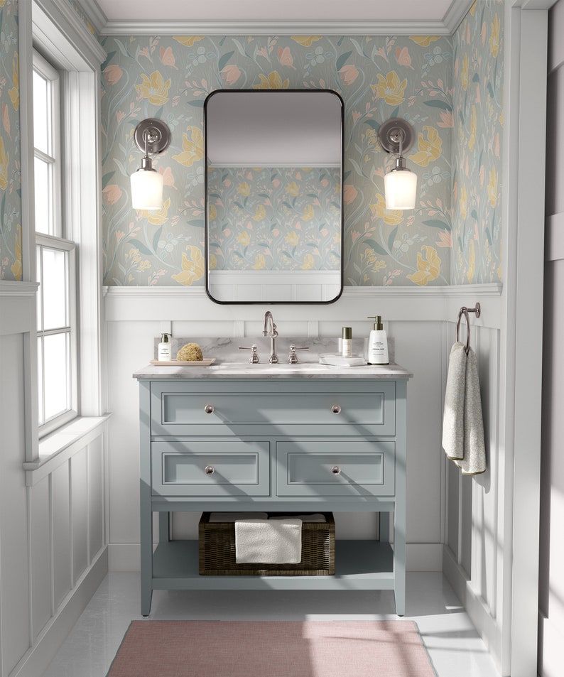 A light-filled bathroom with pastel floral peel and stick wallpaper, a blue vanity, and a pink rug on marble flooring