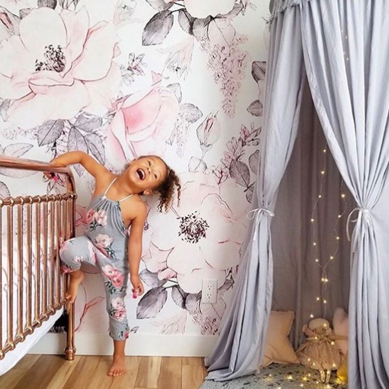 May include: A young girl smiles while standing in a room with a floral wallpaper and a gray canopy tent. The tent has a string of lights inside. The girl is wearing a floral jumpsuit.