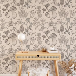 K&ouml;nnte beinhalten: Eine beige Tapete mit einem sich wiederholenden Muster aus schwarz-wei&szlig;en Illustrationen von Pilzen und Pflanzen. Ein Holzschreibtisch mit einer geflochtenen Rattans Schublade steht vor der Tapete. Eine wei&szlig;e Keramiklampe mit einem wei&szlig;en Schirm steht auf dem Schreibtisch. Ein wei&szlig;er Kerzenhalter aus Keramik steht auf dem Schreibtisch. Zwei B&uuml;cher sind auf dem Schreibtisch gestapelt. Ein Hocker aus wei&szlig;em Kunstfell steht vor dem Schreibtisch. Eine Vase mit einem braunen und wei&szlig;en geometrischen Muster steht auf dem Boden. Ein Bund getrockneten beigen Grases steht in der Vase.