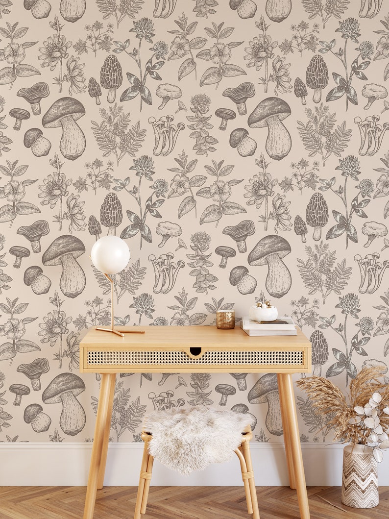 K&ouml;nnte beinhalten: Eine beige Tapete mit einem sich wiederholenden Muster aus schwarz-wei&szlig;en Illustrationen von Pilzen und Pflanzen. Ein Holzschreibtisch mit einer geflochtenen Rattans Schublade steht vor der Tapete. Eine wei&szlig;e Keramiklampe mit einem wei&szlig;en Schirm steht auf dem Schreibtisch. Ein wei&szlig;er Kerzenhalter aus Keramik steht auf dem Schreibtisch. Zwei B&uuml;cher sind auf dem Schreibtisch gestapelt. Ein Hocker aus wei&szlig;em Kunstfell steht vor dem Schreibtisch. Eine Vase mit einem braunen und wei&szlig;en geometrischen Muster steht auf dem Boden. Ein Bund getrockneten beigen Grases steht in der Vase.