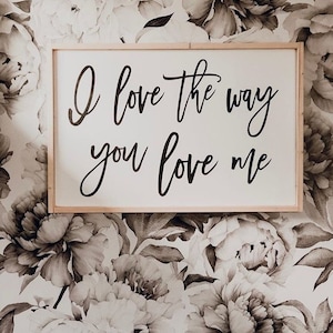 May include: A wooden sign with the text "I love the way you love me" in black cursive lettering against a white background. The sign is hanging on a wall with a floral wallpaper pattern.