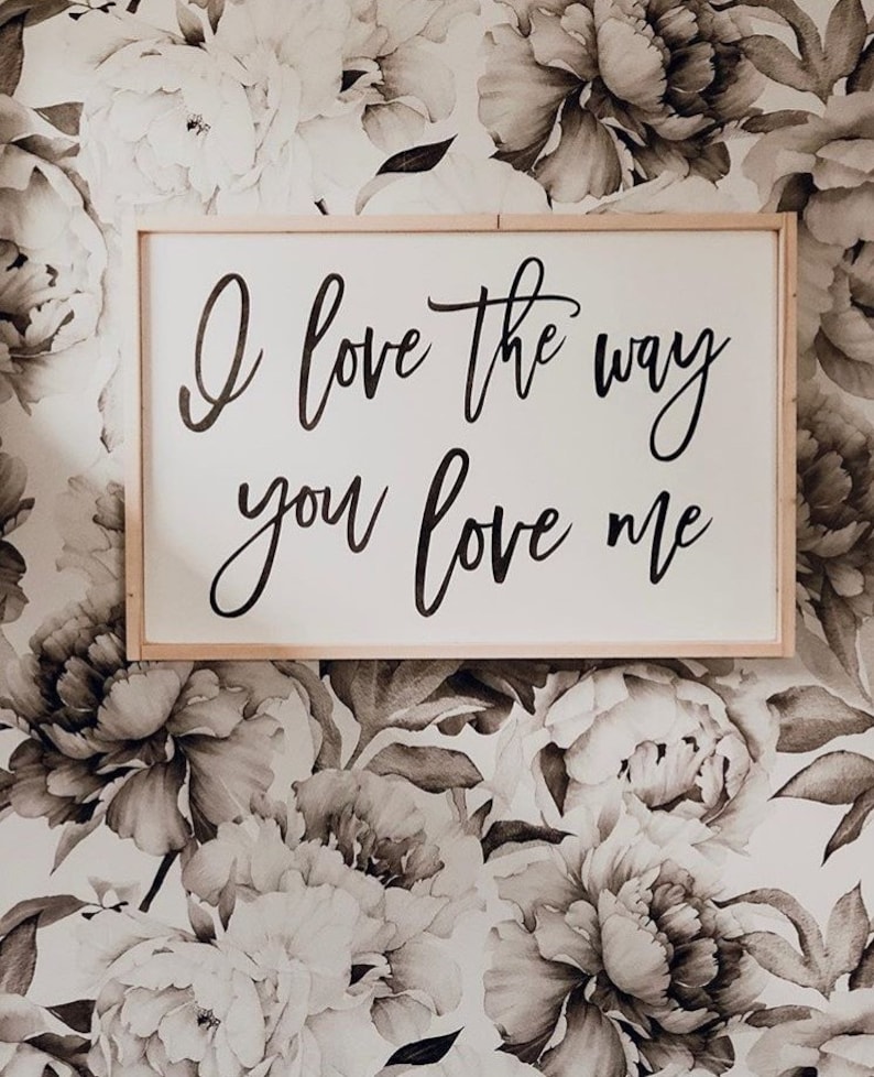 May include: A wooden sign with the text "I love the way you love me" in black cursive lettering against a white background. The sign is hanging on a wall with a floral wallpaper pattern.