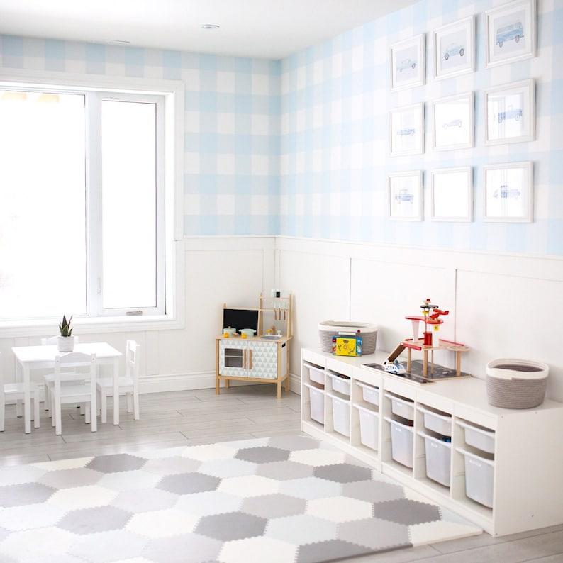 May include: A white playroom with a white table and chairs, a white toy kitchen, a white toy storage unit, and a blue and white checkered wallpaper with framed pictures of cars. The floor is covered with a gray and white hexagonal play mat.