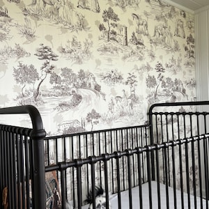 K&ouml;nnte beinhalten: Ein schwarzes Metall-Babybett mit einer wei&szlig;en Matratze und einem wei&szlig;en und schwarzen, gemusterten Kissen. Das Babybett steht vor einer Wand mit einer cremefarbenen Tapete, die ein schwarz-wei&szlig;es Toile-Muster mit B&auml;umen, H&auml;usern und Tieren zeigt.