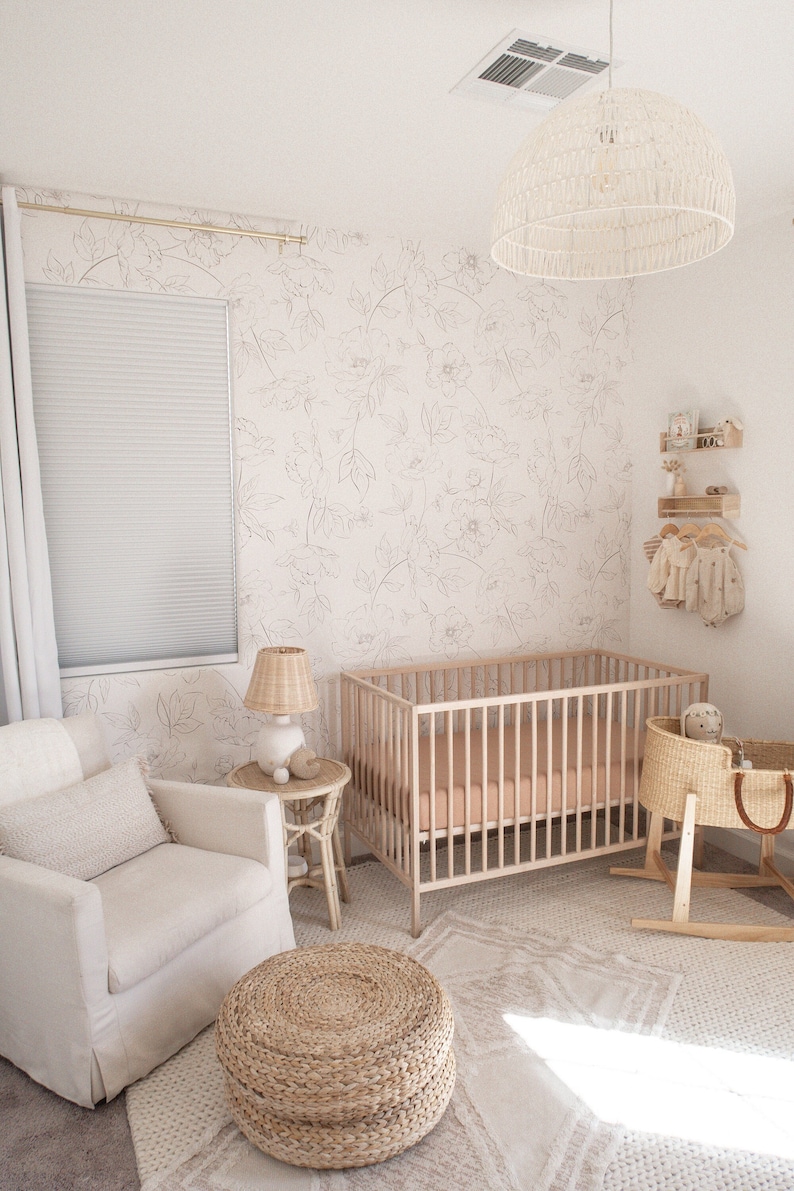 May include: A white crib with a light pink mattress, a white wicker bassinet, a white armchair with a cream pillow, a woven ottoman, and a white lamp with a wicker shade. The room has white walls with a floral wallpaper pattern and a white ceiling fan with a woven shade.