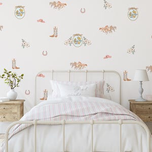 Peut inclure: Une chambre à coucher avec un lit en métal blanc, une couette à rayures et un oreiller avec l'inscription "Good night". Le mur blanc est décoré d'illustrations de chevaux, de bottes de cowboy, de chapeaux, de fleurs et de fers à cheval.