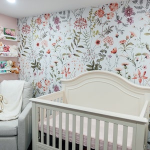 May include: A white cot with a floral patterned wallpaper behind it. The cot has a white mattress and a white headboard. There is a white rocking chair with a white blanket and a stuffed toy in it. The room is decorated with a pink wall and white shelves with stuffed toys on them.