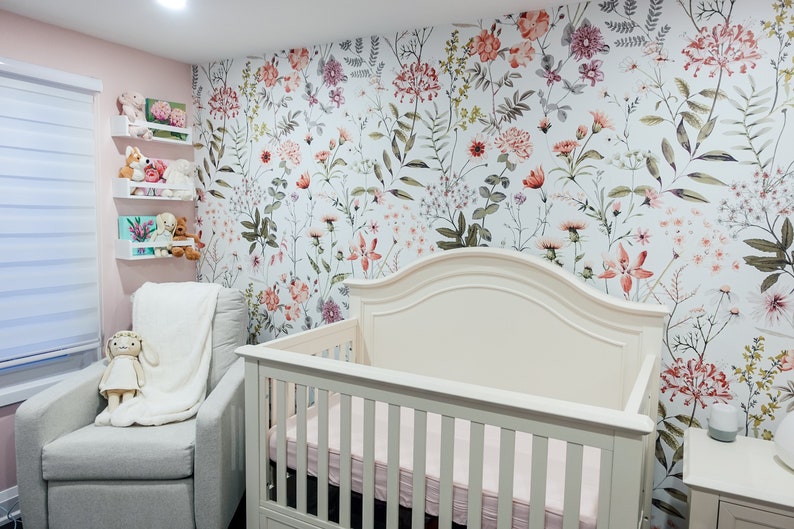 May include: A white cot with a floral patterned wallpaper behind it. The cot has a white mattress and a white headboard. There is a white rocking chair with a white blanket and a stuffed toy in it. The room is decorated with a pink wall and white shelves with stuffed toys on them.
