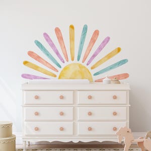 May include: A white dresser with six drawers and rose-colored knobs. Above the dresser is a watercolor sun design with yellow, orange, pink, and blue rays. A rocking horse and decorative boxes are in the foreground.