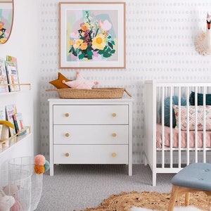 May include: A bright, airy nursery with a white crib, dresser, and a floral art print. The white dresser has three drawers with gold knobs. A woven basket holds star-shaped pillows. A jute rug and a white rug add texture to the room.