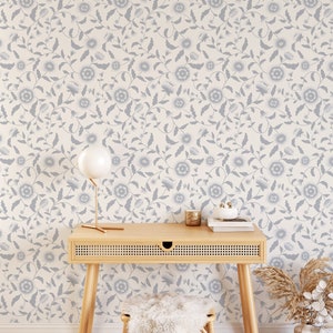 A cozy workspace with blue floral self-adhesive, removable wallpaper. A stylish wooden desk with angled legs holds a lamp, books, and small gold accessories. A fluffy stool and a vase with dried plants add warmth to the space