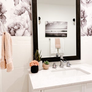 May include: A white bathroom vanity with a white countertop and a white porcelain sink. The vanity has a single drawer with a brushed nickel handle. There are two chrome faucets and a soap dispenser on the countertop. There are two potted plants on the countertop, one with green leaves and the other with pink flowers. A large framed mirror hangs above the vanity. The mirror reflects a black and white landscape painting and two towels hanging on a towel rack.