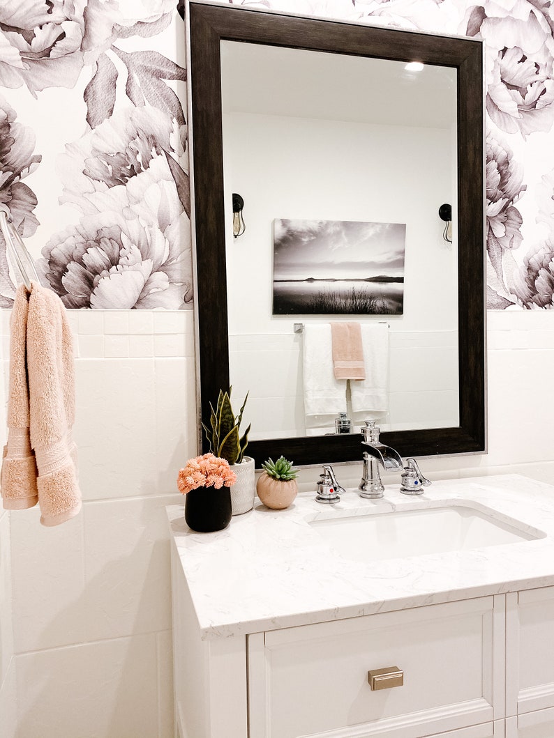 May include: A white bathroom vanity with a white countertop and a white porcelain sink. The vanity has a single drawer with a brushed nickel handle. There are two chrome faucets and a soap dispenser on the countertop. There are two potted plants on the countertop, one with green leaves and the other with pink flowers. A large framed mirror hangs above the vanity. The mirror reflects a black and white landscape painting and two towels hanging on a towel rack.