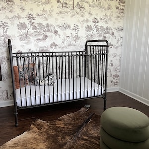 K&ouml;nnte beinhalten: Ein schwarzes Metall-Babybett mit einer wei&szlig;en Matratze und einem braunen Kuhfellteppich. Ein gr&uuml;ner Samt-Hocker steht in der Ecke des Raumes.