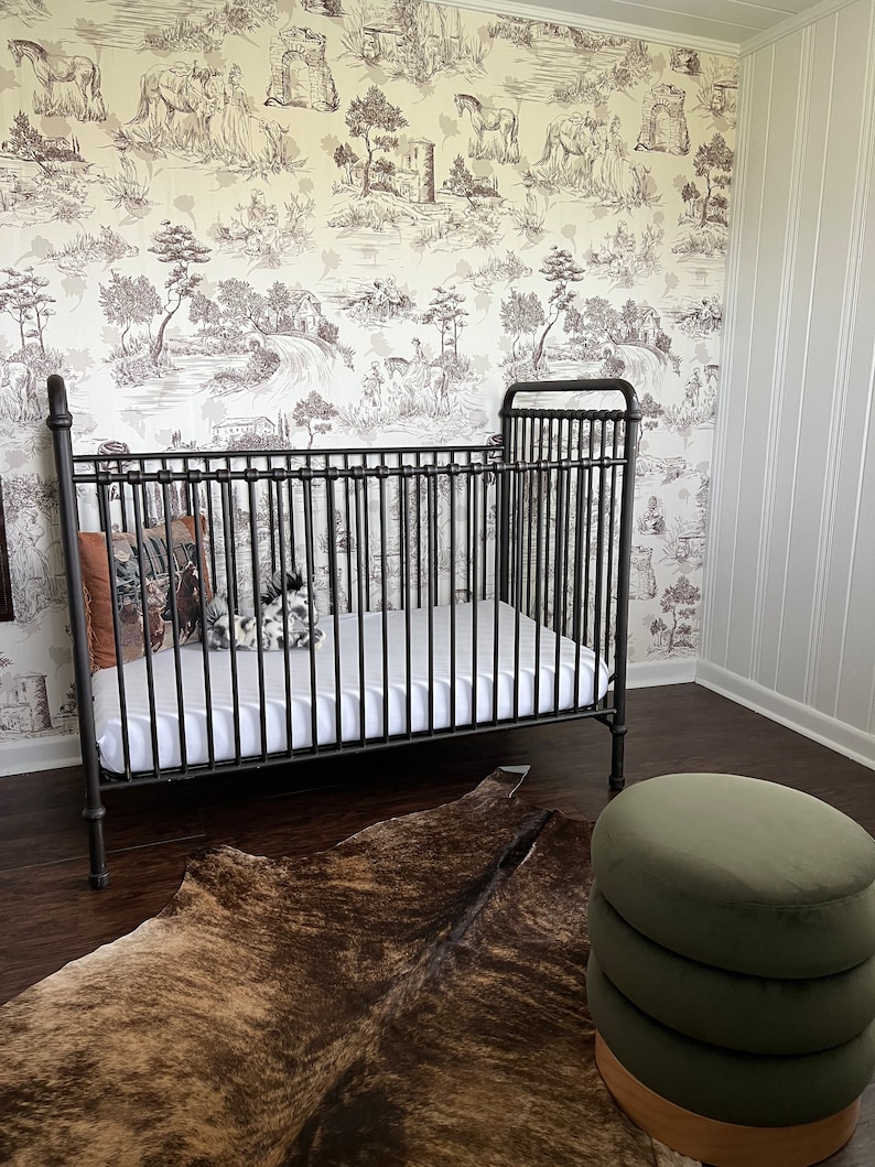 K&ouml;nnte beinhalten: Ein schwarzes Metall-Babybett mit einer wei&szlig;en Matratze und einem braunen Kuhfellteppich. Ein gr&uuml;ner Samt-Hocker steht in der Ecke des Raumes.