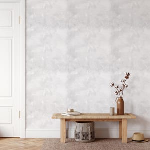 May include: An interior scene with a light wood bench, a woven top, a decorative basket, and a brown vase with cotton stems. A straw hat rests on the floor. The wall has a textured, light grey finish. The room has a neutral colour palette.