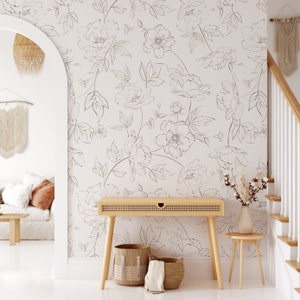 May include: A white wall with a black and white floral wallpaper pattern. A wooden console table with two woven baskets underneath. A small wooden side table with a vase of dried flowers.