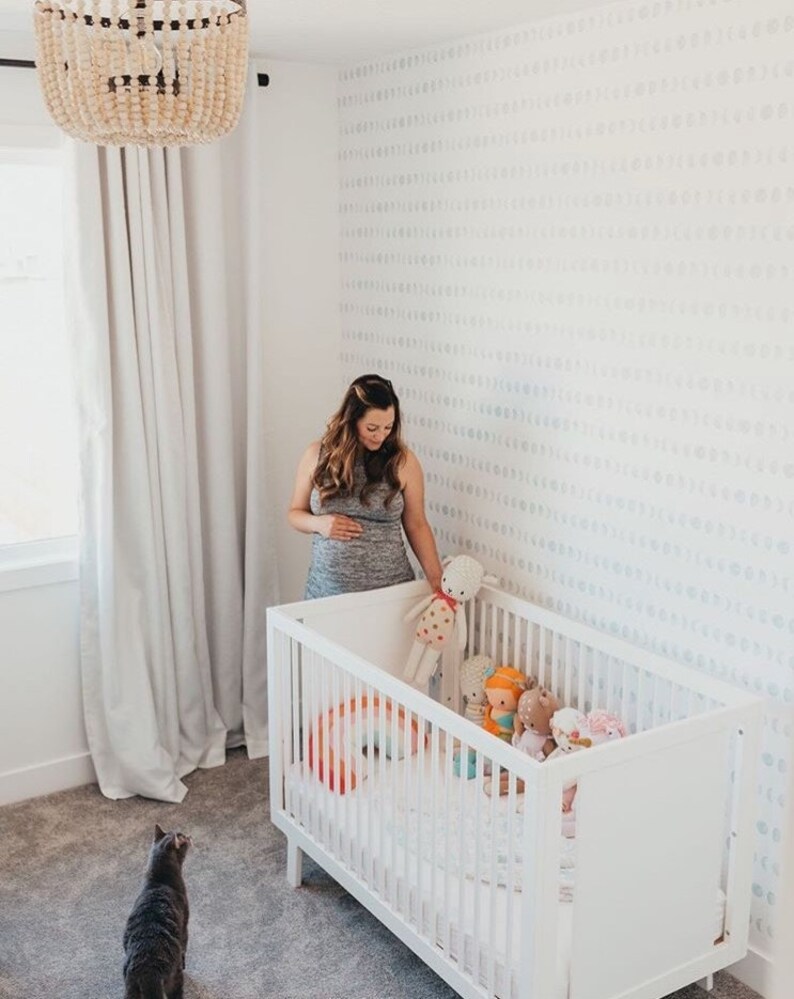 May include: A white crib filled with stuffed animals and a colorful pillow. The crib is in a nursery with a patterned accent wall and a beaded chandelier. A person stands next to the crib, and a gray cat is in the foreground.
