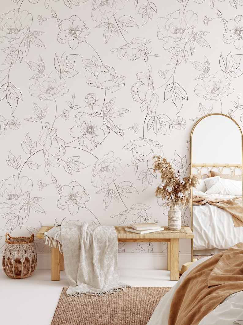 May include: A white bedroom with a floral wallpaper featuring a delicate line drawing of flowers. A wooden bench with a white throw blanket sits in front of the wallpaper. A wicker basket sits on the floor next to the bench. A mirror with a gold frame is on the right side of the image. A bed with a brown throw blanket is in the foreground.