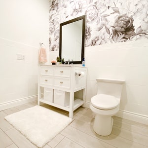May include: A white bathroom with a white toilet, a white vanity with drawers and shelves, a white rug, and a floral wallpaper with black and white peonies.
