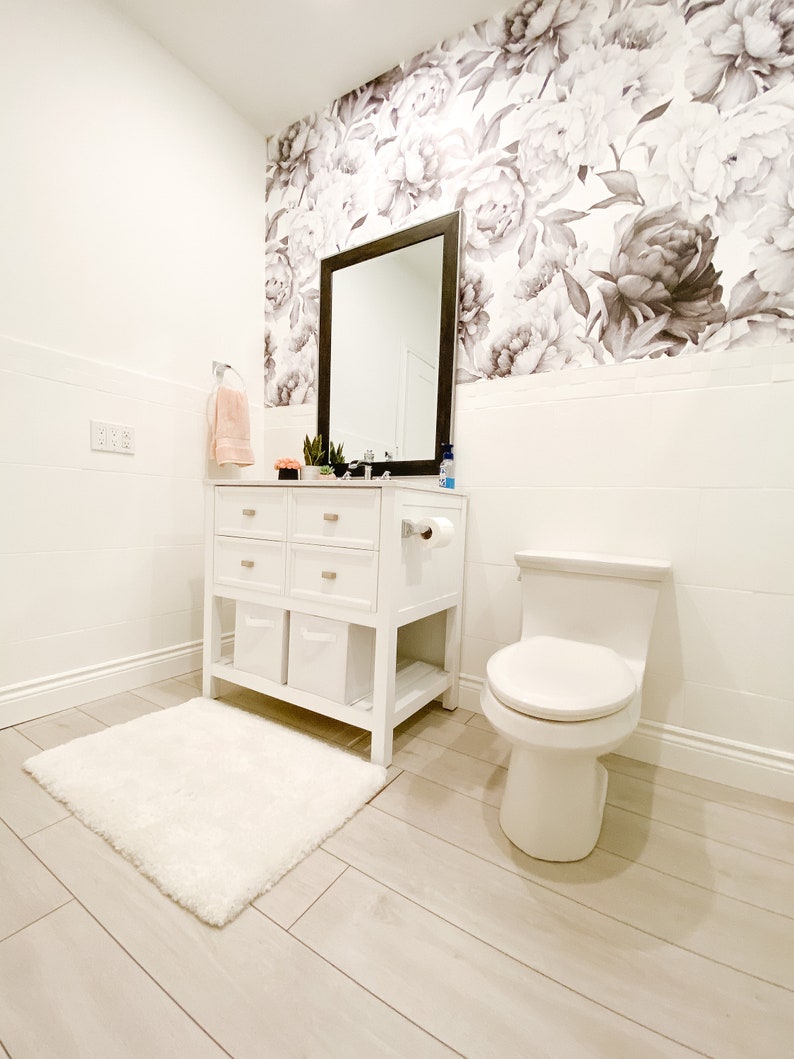 May include: A white bathroom with a white toilet, a white vanity with drawers and shelves, a white rug, and a floral wallpaper with black and white peonies.