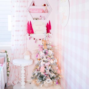 May include: A flocked Christmas tree decorated with pink and white ornaments, gingerbread figures, and unicorn motifs. Above the tree, a white hexagonal shelf holds small holiday decor, and a shelf below displays pink bottle brush trees.
