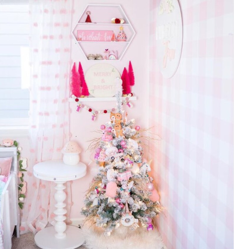 May include: A flocked Christmas tree decorated with pink and white ornaments, gingerbread figures, and unicorn motifs. Above the tree, a white hexagonal shelf holds small holiday decor, and a shelf below displays pink bottle brush trees.