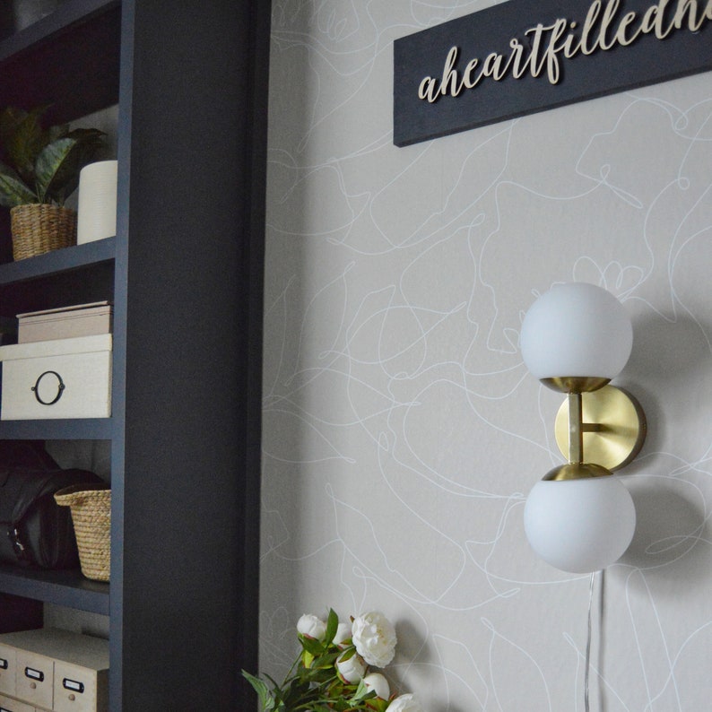 May include: A gold wall sconce with two white globe light fixtures. The sconce is mounted on a light gray wall with a white abstract pattern. The text "a heart filled" is visible on a black sign above the sconce.