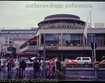May include: A vintage photograph of a street scene in a European city. A large building with a sign that reads "KRANZHORN" is visible in the background. People are walking on the sidewalk in front of the building. There are cars parked on the street.
