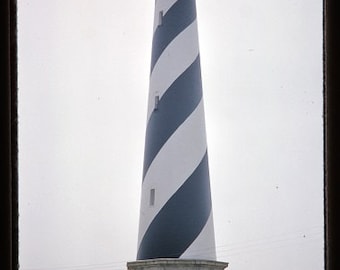 May include: A black and white striped lighthouse with a brick base against a cloudy sky. The lighthouse is tall and slender with a small lantern room at the top.