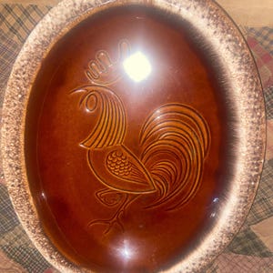 May include: Oval-shaped, brown ceramic serving platter with a raised rooster design in the center. The rooster is depicted in a stylized, vintage style. The rim of the platter has a speckled, cream-colored border.