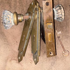 May include: Antique door hardware set featuring two glass doorknobs, brass plates, a door latch, and a skeleton key. The doorknobs have a ribbed design. The brass components show signs of age and wear, adding to their vintage appeal.