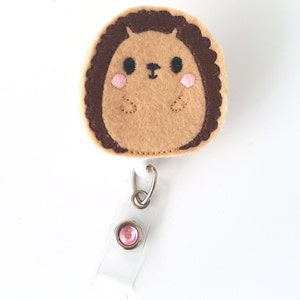 Könnte beinhalten: Ein brauner und beiger Filz-Igel mit einem rosafarbenen Strass-Badgehalter. Der Igel hat ein süßes gesticktes Gesicht mit schwarzen Augen und einer rosa Nase.