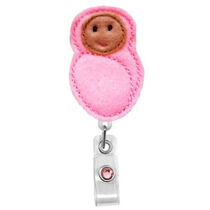 Puede incluir: Muñeca de fieltro rosa con cara marrón y un strass rosa en un portagafete retráctil.