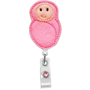 Puede incluir: Un portatarjetas de bebé de fieltro rosa con una cara sonriente y una gema rosa. El portatarjetas tiene un cordón retráctil blanco y un clip de plástico transparente.