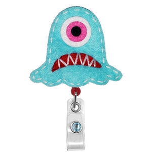 Könnte beinhalten: Ein blaues Filzmonster mit einem einzigen rosa Auge und roten gestickten Zähnen. Das Monster ist an einem durchsichtigen Plastik-Badgehalter mit einem blauen Strassstein befestigt.