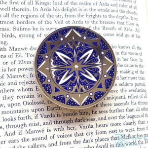 Puede incluir: Un pin redondo de esmalte dorado con un diseño detallado de estrella azul y blanca. El pin tiene un borde dorado y patrones intrincados, probablemente inspirados en temas celestiales o de fantasía. El pin está sobre un libro.