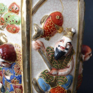 Może przedstawiać: Biały ceramiczny wazon z złotymi akcentami i wielokolorowym wzorem. Wazon przedstawia czerwoną rybę, mężczyznę z brodą i wz&oacute;r kwiatowy. Wazon jest ozdobiony złotą obw&oacute;dką i ma fakturowaną powierzchnię.