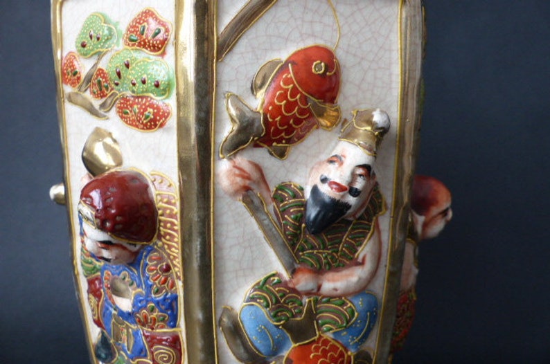 Może przedstawiać: Biały ceramiczny wazon z złotymi akcentami i wielokolorowym wzorem. Wazon przedstawia czerwoną rybę, mężczyznę z brodą i wz&oacute;r kwiatowy. Wazon jest ozdobiony złotą obw&oacute;dką i ma fakturowaną powierzchnię.