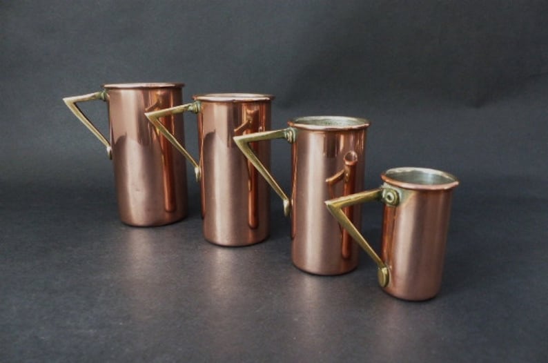 May include: Four copper measuring cups with brass handles. The cups are nested together, with the largest cup at the bottom and the smallest cup at the top.