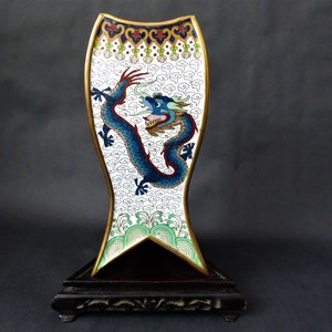 K&ouml;nnte beinhalten: Eine wei&szlig;e Cloisonn&eacute;-Emailvase mit einem blauen Drachenmotiv auf einem schwarzen Holzsockel. Die Vase ist fischf&ouml;rmig und hat einen goldenen Rand.