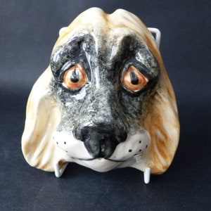 Puede incluir: Una escultura de cerámica para colgar en la pared que representa la cabeza de un perro. El perro tiene pelo marrón y negro, con la nariz negra y los ojos marrones. La escultura está pintada con un estilo realista.