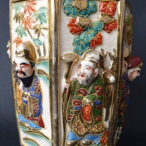 Może przedstawiać: Biały ceramiczny wazon o sześciokątnym kształcie z pozłacanymi zdobieniami. Wazon jest ozdobiony kolorowymi obrazami ludzi i kwiat&oacute;w. Wazon jest tradycyjnym japońskim wzorem.