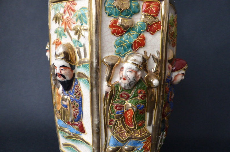 Może przedstawiać: Biały ceramiczny wazon o sześciokątnym kształcie z pozłacanymi zdobieniami. Wazon jest ozdobiony kolorowymi obrazami ludzi i kwiat&oacute;w. Wazon jest tradycyjnym japońskim wzorem.