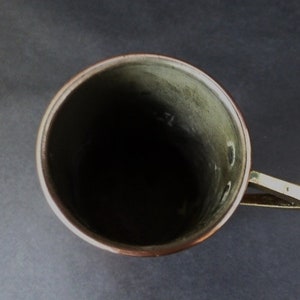 May include: A copper mug with a brass handle. The mug is empty and has a green patina inside.