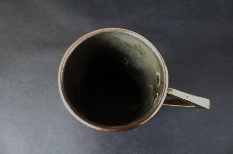 May include: A copper mug with a brass handle. The mug is empty and has a green patina inside.