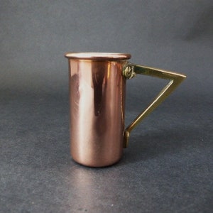 May include: A copper measuring cup with a brass handle. The cup is cylindrical and has a rounded bottom. The handle is attached to the side of the cup and has a curved shape.