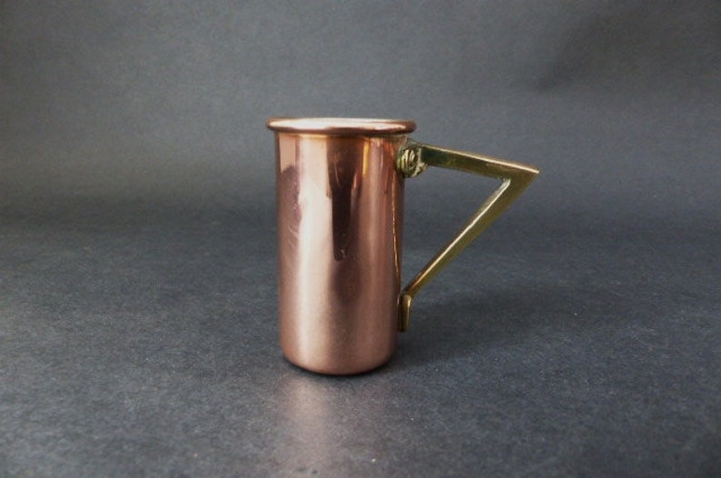 May include: A copper measuring cup with a brass handle. The cup is cylindrical and has a rounded bottom. The handle is attached to the side of the cup and has a curved shape.